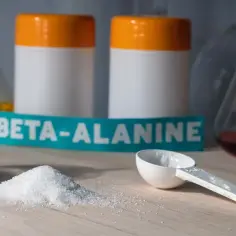 Biały proszek beta-alaniny na blacie, obok miarki i buteleczek laboratoryjnych. Uważaj na potencjalne beta-alanina skutki uboczne.
