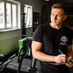 Trener Paweł notuje najlepsze ćwiczenia na biceps. W tle siłownia z bieżniami i oknami.