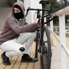 Mężczyzna w masce i kapturze próbuje dokonać kradzieży roweru, rozpinając zamek.