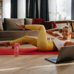 Uśmiechnięta kobieta wykonuje ćwiczenia w domu na macie, patrząc na laptopa.