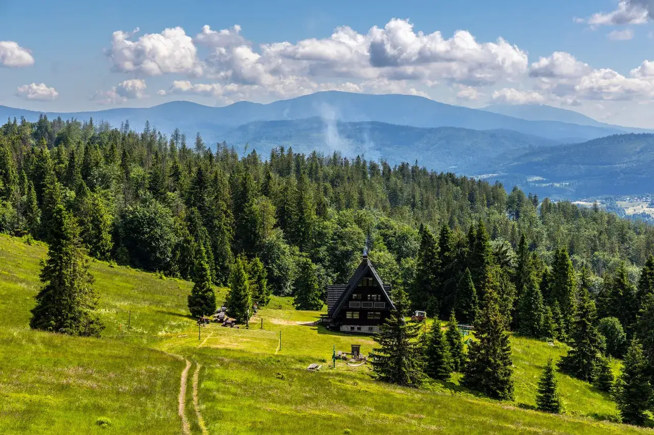 Widok na beskid żywiecki szczyty z chatką w lesie. Dym unosi się z komina, a chmury na niebie dodają uroku krajobrazowi.