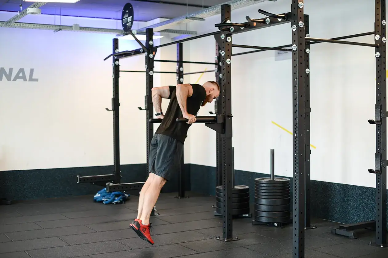 Mężczyzna wykonuje ćwiczenie dipy na triceps na siłowni. W tle stojaki i obciążenia.
