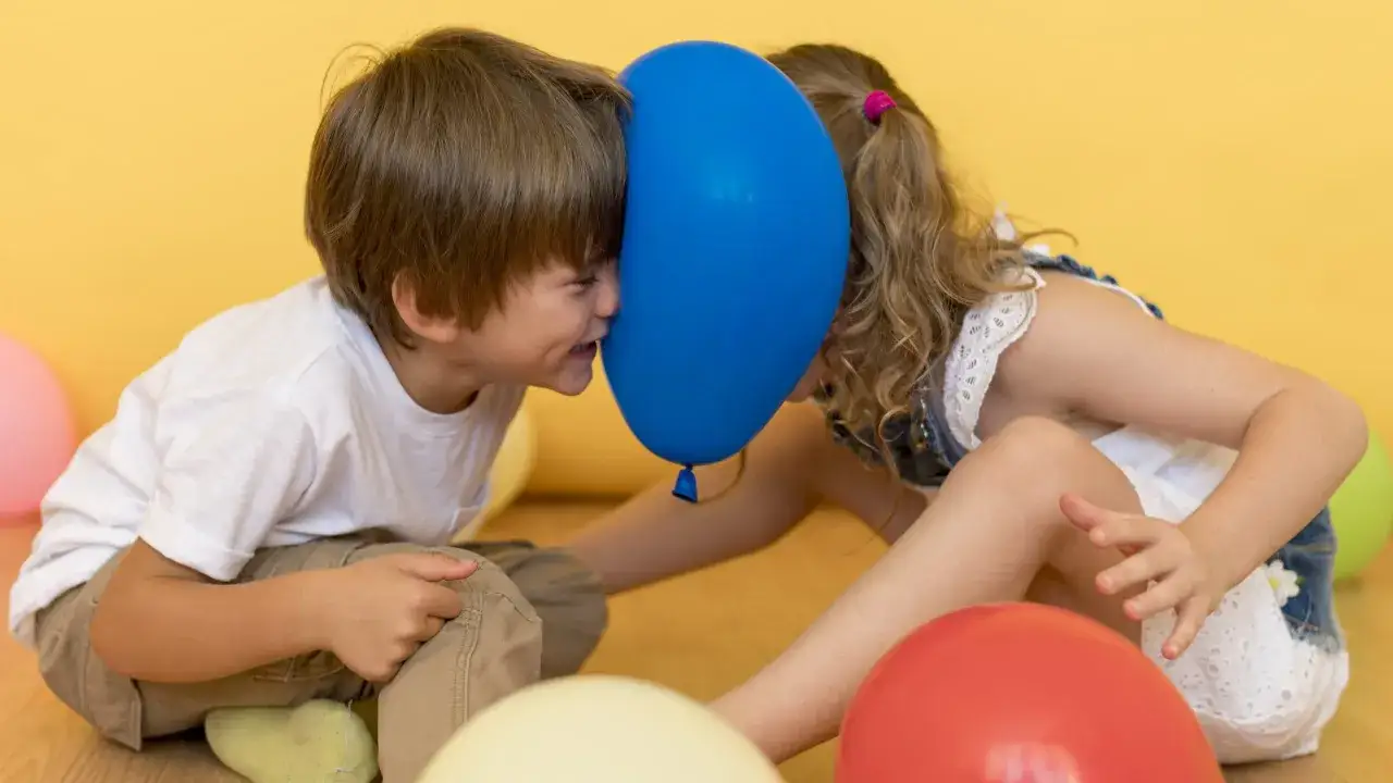 Dzieci bawią się balonem, co stanowi świetne ćwiczenia sensoryczne. Chłopiec i dziewczynka zbliżają twarze do niebieskiego balonu, śmiejąc się.