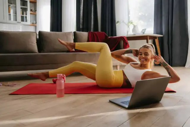 Uśmiechnięta kobieta wykonuje ćwiczenia w domu na macie, patrząc na laptopa.